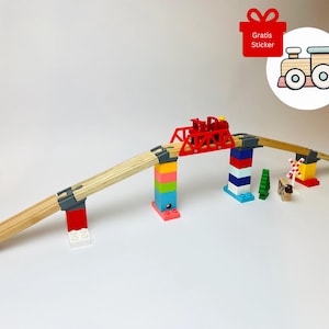 Op de afbeelding: Een houten speelgoedtreinset met een rode brug en kleurrijke steunblokken. De set bevat een rode trein, groene bomen en een kleine dierfiguur. Een sticker met een treinillustratie en de tekst "Gratis Sticker" is ook zichtbaar.