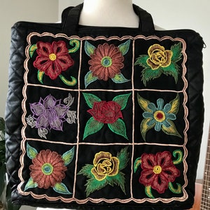 May include: Black quilted tote bag with a zipper closure and two top handles. The bag features a patchwork design with nine embroidered floral motifs in various colors, including red, yellow, green, blue, and purple.