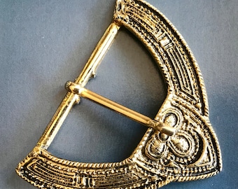 May include: A gold-toned metal belt buckle with an intricate, ornate design. The buckle is shaped like a crescent moon and has a detailed pattern of lines and swirls.