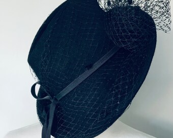 Puede incluir: Un sombrero de fieltro negro con velo de red negro y un lazo de cinta negro.