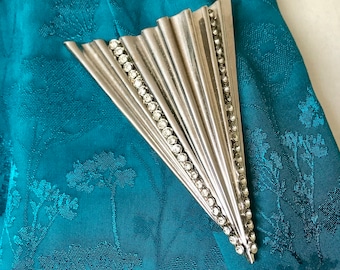 May include: A silver-toned brooch in the shape of a fan with clear rhinestones around the edges. The brooch is on a teal fabric background.