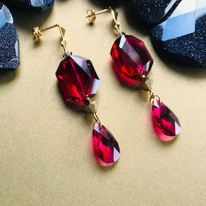 May include: A pair of dangle earrings featuring red crystal teardrop and octagon shaped beads. The earrings are made with gold tone metal.