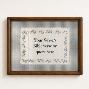 May include: A rectangular dark wood frame containing a white fabric insert with a blue floral border and scalloped edges. The center of the fabric features the black printed text: Your favorite Bible verse or quote here, set against a blue gingham mat.