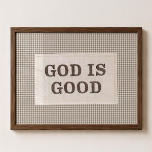 Puede incluir: Un cuadro rectangular con marco de madera marrón oscuro. En el centro, un parche de tela color crema muestra el texto bordado en marrón oscuro "GOD IS GOOD", sobre un fondo con estampado de pata de gallo en beige y blanco.