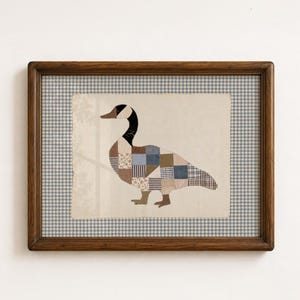 Puede incluir: Una obra de arte enmarcada que presenta una ilustración de un ganso de patchwork compuesto por varios cuadrados de tela en tonos azul, marrón y crema. El ganso tiene el cuello y la cabeza negros, sobre un fondo crema con un marco de madera.