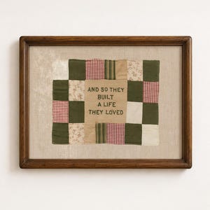 May include: A framed textile wall art piece featuring a patchwork quilt design in shades of olive green, cream, and red gingham. The centre panel is embroidered with the text AND SO THEY BUILT A LIFE THEY LOVED in dark green thread.