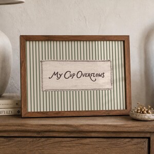 May include: A rectangular wooden picture frame containing a light-coloured fabric insert with the text "My Cup Overflows" in dark brown script. The background of the insert features thin vertical green stripes and a decorative stitched border.