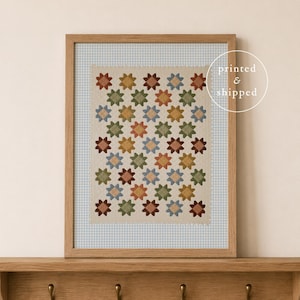 May include: A framed print featuring a repeating pattern of multi-colored star shapes in shades of rust, olive green, and light blue on a cream background, set within a light blue gingham border, displayed in a light wood frame on a wooden shelf.