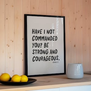 K&ouml;nnte beinhalten: Ein schwarzer gerahmter Druck mit wei&szlig;em Hintergrund und schwarzem Text, der "Have I not commanded you? Be strong and courageous. Joshua 1:9" lautet. Der Druck h&auml;ngt an einer hellbraunen Holzwand.