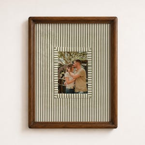 May include: A rectangular dark brown wooden picture frame containing a photograph of two people embracing. The frame features a mat with a vertical white and sage green pinstripe pattern, surrounding the central portrait of the couple.