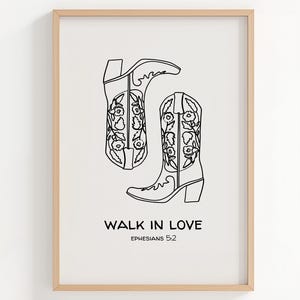 May include: A framed print with a black line drawing of two cowboy boots with floral patterns. The text "WALK IN LOVE" and "EPHESIANS 5:2" are below the boots. The frame is a light wood colour.