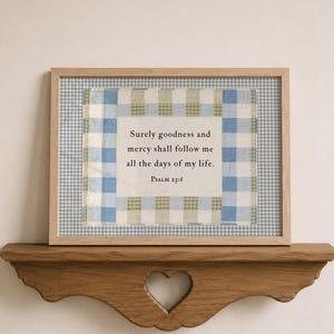 May include: A rectangular wooden frame containing a fabric print with a blue and white gingham border. The center features the text: Surely goodness and mercy shall follow me all the days of my life. Psalm 23:6, printed on a light cream background.