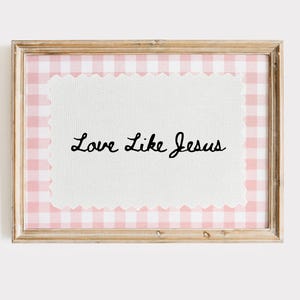 May include: A framed print with the text "Love Like Jesus" in elegant script. The print features a white, scalloped-edge rectangle set against a pink and white gingham pattern. The frame is made of light-colored wood.