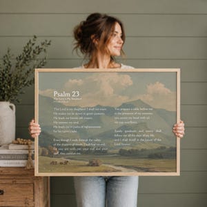 May include: A framed print of Psalm 23, displayed against a landscape painting. The text is in a classic serif font. The artwork has a light wood frame. The print is held by a person.