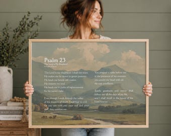 Pode incluir: Uma impressão emoldurada com o Salmo 23, com uma pintura de paisagem ao fundo. O texto está em uma fonte serif clássica. A obra de arte tem uma moldura de madeira clara e é segurada por uma pessoa.