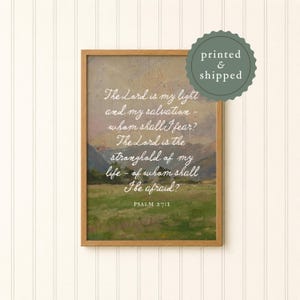 May include: A framed print featuring a landscape painting with a quote from Psalm 27:1 in white script. The artwork has a green and brown color palette, with a mountain range in the background. A green circular sticker says "printed & shipped".