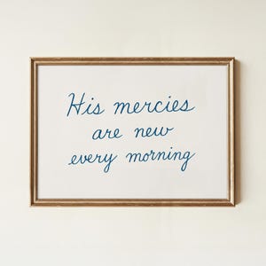Op de afbeelding: Een ingelijste print met de tekst "His mercies are new every morning" in een sierlijk lettertype. De tekst is donkerblauw op een witte achtergrond. De lijst is goudkleurig.