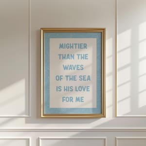 May include: A framed print with the words "Mightier Than The Waves Of The Sea Is His Love For Me" in light blue text. The text is centered on a cream-colored background with a light blue border, set within a gold frame. The print hangs on a white wall.