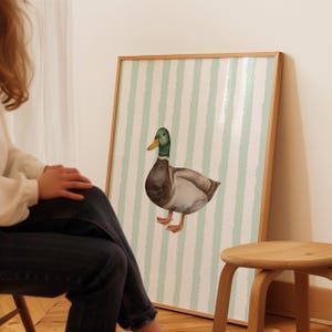 Op de afbeelding: Een aquarel van een wilde eend op een lichtgroen-wit gestreepte achtergrond. De eend kijkt naar links en heeft een groene kop, een bruin lichaam en grijze vleugels.