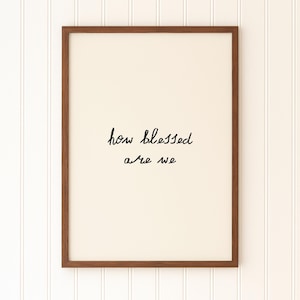 May include: A framed print with the words "how blessed are we" in black script. The print has a cream-colored background and is encased in a dark brown wooden frame. The background is a white vertical shiplap wall.