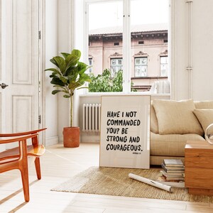 K&ouml;nnte beinhalten: Ein beigefarbener gerahmter Poster mit schwarzem Text, der "Have I not commanded you? Be strong and courageous. Joshua 1:7" lautet, h&auml;ngt an einer wei&szlig;en Wand in einem Wohnzimmer mit einem braunen Sofa, einem Holz-Couchtisch und einer Topfpflanze.