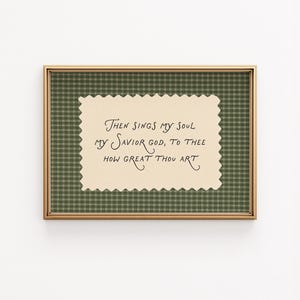 May include: A framed print with a gold-colored frame. The print features a cream-colored, scalloped-edge paper with handwritten text in black ink. The background is a green plaid pattern. The text reads "Then sings my soul my Savior God, to Thee how great thou art."
