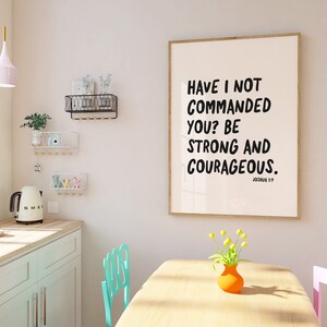 K&ouml;nnte beinhalten: Ein gerahmter Druck mit wei&szlig;em Hintergrund und schwarzem Text, der "Have I not commanded you? Be strong and courageous." lautet. Der Text ist in fetter, handschriftlicher Schrift. Der Rahmen ist in hellem Holzton. Der Druck h&auml;ngt an einer wei&szlig;en Wand in einem Raum mit einem Tisch aus hellem Holz und zwei St&uuml;hlen. Auf dem Tisch steht eine Vase mit gelben Blumen.