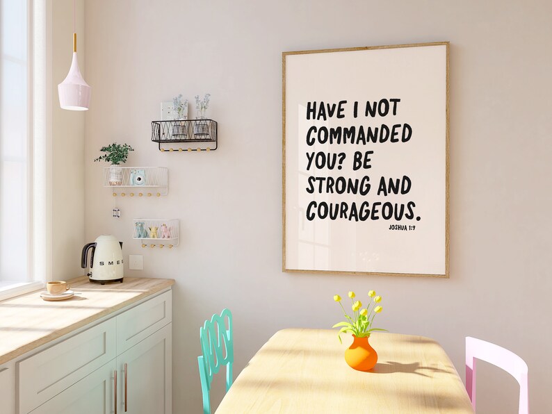K&ouml;nnte beinhalten: Ein gerahmter Druck mit wei&szlig;em Hintergrund und schwarzem Text, der "Have I not commanded you? Be strong and courageous." lautet. Der Text ist in fetter, handschriftlicher Schrift. Der Rahmen ist in hellem Holzton. Der Druck h&auml;ngt an einer wei&szlig;en Wand in einem Raum mit einem Tisch aus hellem Holz und zwei St&uuml;hlen. Auf dem Tisch steht eine Vase mit gelben Blumen.