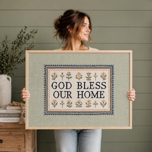 Pode incluir: Um mostruário de ponto cruz retangular emoldurado com o texto "GOD BLESS OUR HOME" em letras azul-escuras. O design inclui uma borda de pequenos motivos florais em tons suaves de rosa, verde e castanho sobre um fundo creme claro.