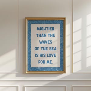 Op de afbeelding: Een ingelijste print met de tekst "Machtiger dan de golven van de zee is zijn liefde voor mij." De blauwe tekst staat op een witte achtergrond, met een blauwe patroonrand en een gouden lijst. De print hangt aan een witte muur.