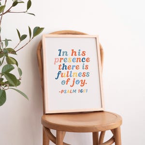 Puede incluir: Una impresión enmarcada con las palabras "In his presence there is fullness of joy" en texto colorido. La impresión está en un marco de madera clara y está apoyada en una silla de madera.