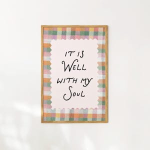 Op de afbeelding: Een ingelijste print met de tekst "IT IS WELL WITH MY SOUL" in zwart schrift. De print heeft een roze achtergrond met een geschulpte rand, omgeven door een kleurrijk ruitpatroon in groen, oranje en roze.