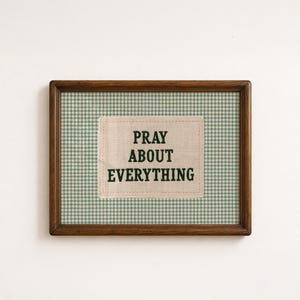 May include: A rectangular wooden frame containing a green and white houndstooth patterned background. Centered is a rectangular fabric patch with the dark green embroidered text PRAY ABOUT EVERYTHING, featuring visible stitching around the edges.