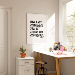 K&ouml;nnte beinhalten: Ein wei&szlig; gerahmter inspirierender Zitatdruck mit schwarzem Text, der "Have I not commanded you? Be strong and courageous." lautet. Der Druck h&auml;ngt an einer wei&szlig;en Wand &uuml;ber einem wei&szlig;en Schreibtisch mit einem Holzrahmen. Der Schreibtisch hat einen Laptop, Kopfh&ouml;rer und andere B&uuml;roartikel.