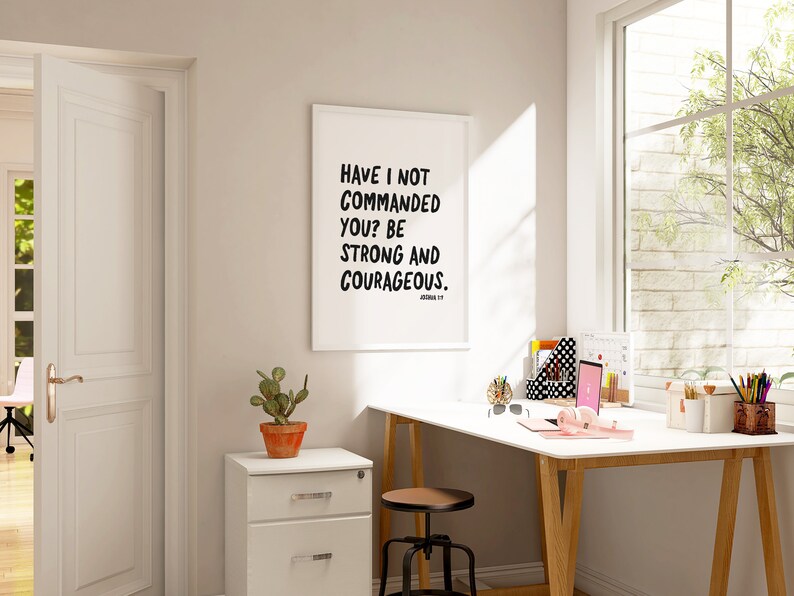 K&ouml;nnte beinhalten: Ein wei&szlig; gerahmter inspirierender Zitatdruck mit schwarzem Text, der "Have I not commanded you? Be strong and courageous." lautet. Der Druck h&auml;ngt an einer wei&szlig;en Wand &uuml;ber einem wei&szlig;en Schreibtisch mit einem Holzrahmen. Der Schreibtisch hat einen Laptop, Kopfh&ouml;rer und andere B&uuml;roartikel.