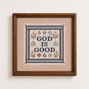 May include: A square cross-stitch embroidery in a dark wood frame featuring the text GOD IS GOOD in dark blue letters. The design includes small floral and botanical motifs in muted tones of pink, green, and blue on a cream fabric background.