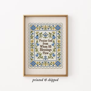 May include: A framed cross-stitch embroidery featuring the text Praise God from Whom All Blessings Flow in brown lettering. The design is surrounded by a floral border of blue bell-shaped flowers and small yellow blossoms, all enclosed in a gold frame.