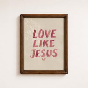 Puede incluir: Un marco de madera rectangular que contiene una impresión de color crema con el texto LOVE LIKE JESUS escrito en un estilo de pincelada de color rojo rosado. Debajo del texto hay un pequeño símbolo de corazón pintado a mano, centrado.