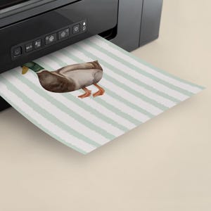 Op de afbeelding: Een printer print een vel papier met een aquarel-illustratie van een eend op een lichtblauwe en witte gestreepte achtergrond.