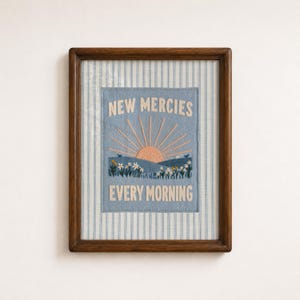 May include: A framed embroidery art piece featuring a light blue fabric background with white vertical stripes. The center displays a stitched sunrise over rolling hills and flowers with the text NEW MERCIES EVERY MORNING in cream thread.