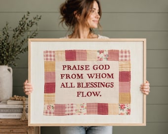 Könnte beinhalten: Ein gerahmtes Textil-Wandbild mit einem zentralen cremefarbenen Stoffpaneel und dem gestickten dunkelroten Text "PRAISE GOD FROM WHOM ALL BLESSINGS FLOW." Das Paneel ist von einem Patchwork-Rahmen aus rosa, roten und geblümten Quadraten umgeben.