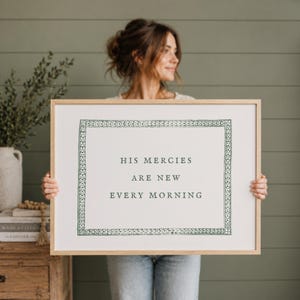 Könnte beinhalten: Ein gerahmter Druck mit dem Text "HIS MERCIES ARE NEW EVERY MORNING" in einer Serifenschrift. Der Druck hat einen dekorativen Rand und wird von einer Person gehalten. Der Rahmen hat eine helle Holzfarbe.