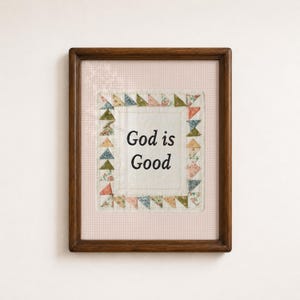 May include: A framed wall art piece featuring the text God is Good in black lettering on a white fabric centre. The centre is surrounded by a border of small, multi-coloured floral fabric triangles, all set against a light pink gingham background.