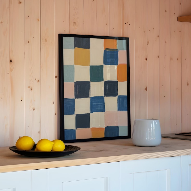 May include: A framed abstract painting with a checkered pattern in shades of blue, pink, yellow, and white. The painting is hanging on a light wood wall. Three yellow lemons are in a black bowl on a white countertop.