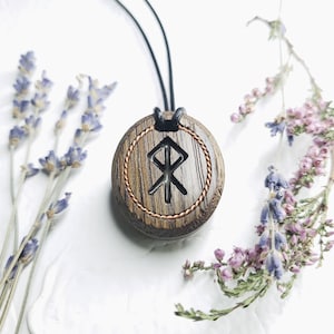 Könnte beinhalten: Halskette mit Holzanhänger und geschnitztem Runensymbol, umgeben von kupferfarbenem Draht und einem geflochtenen Rand. Der Anhänger hängt an einer schwarzen Kordel. Lavendel- und violette Blumen sind um den Anhänger angeordnet.