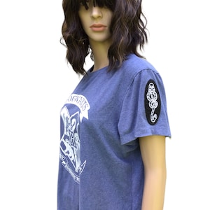 以下が含まれることがあります： 右袖に黒い刺繍の死の秘宝のシンボルパッチが付いた、白いホグワーツの紋章のグラフィックが施された青いTシャツです。