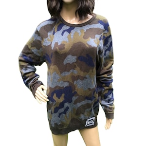 May include: A long-sleeved camouflage sweater with a blue, green, and brown pattern. The sweater has a crew neck and a small patch on the bottom right side that says "Rock".