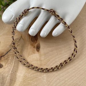 May include: A braided bracelet with brown and gold threads. The bracelet is displayed on a white hand-shaped display against a wooden background. The bracelet's design features a woven pattern.