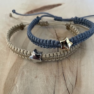 以下が含まれることがあります： 星のチャームが付いた2つのマクラメブレスレット。1つのブレスレットは薄いタン色で、もう1つはくすんだ青色です。星のチャームはシルバーとゴールドです。ブレスレットはスライドノット留めで調節可能です。