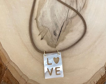Könnte beinhalten: Halskette mit rechteckigem Anhänger, in dem das Wort "LOVE" ausgeschnitten ist, mit einem herzförmigen Ausschnitt über dem "VE". Der perlmuttfarbene Anhänger hängt an einer braunen Kordelkette mit goldfarbenen Verschlüssen.